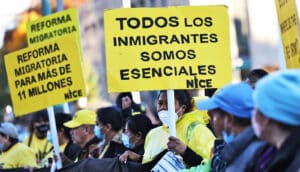 people in crowd hold yellow signs that say "todos los inmigrantes somos esenciales NICE" and "reforma migratoria para más 11 millones"