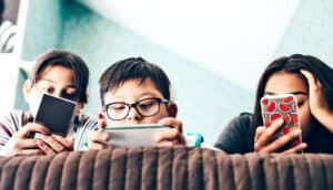 Three kids lay on a bed using smartphones.