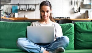 A woman sits on a green couch with her laptop on her folded legs.