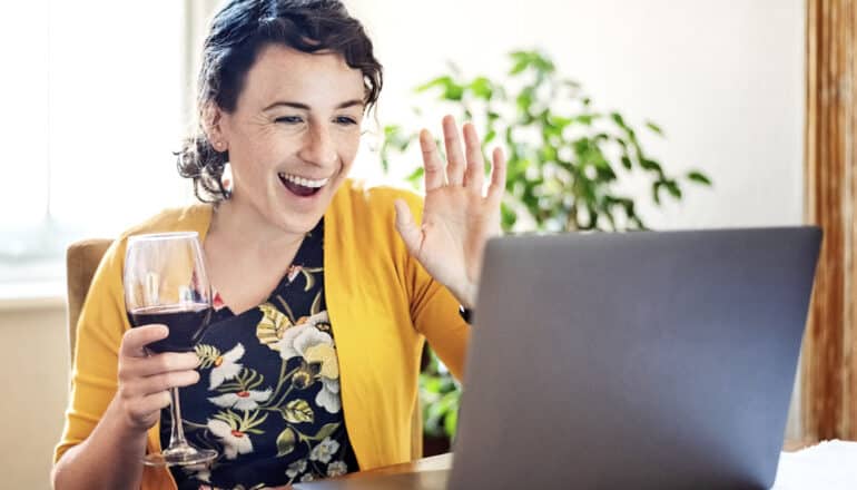 person holding glass of red wine waves at laptop screen