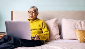 An older woman in a yellow sweater sits on her bed with her laptop.