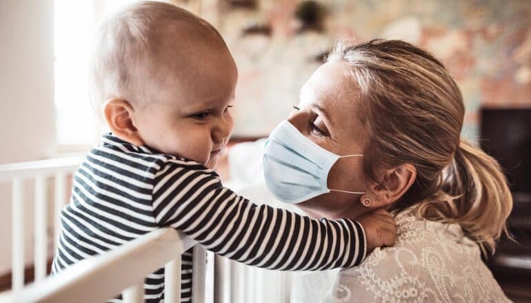 A woman wearing a face mask leans down to a baby standing in a crib.