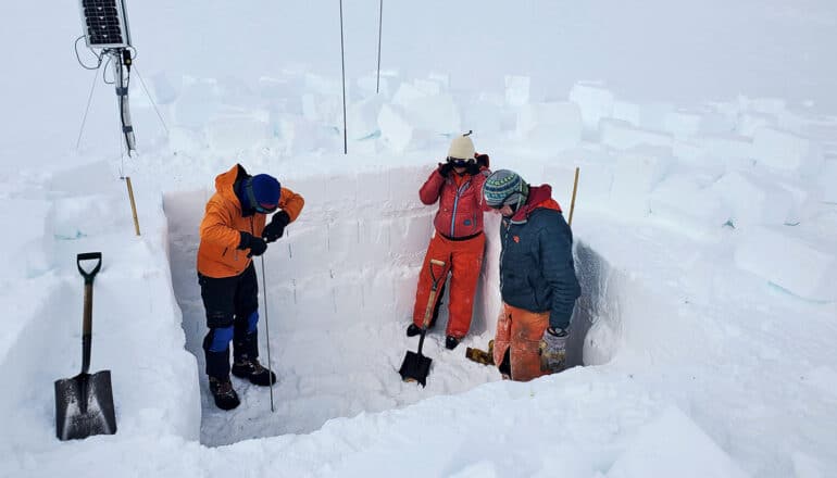 A group of researchers in a square hole dug into ice and snow, with cube-like chunks lifted out and thrown to the side.