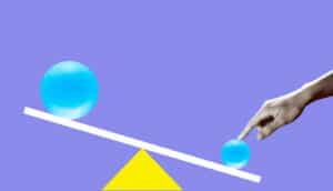 Black and white hand pushes down small blue ball on balance scale, larger blue ball on opposite high side of scale, purple background