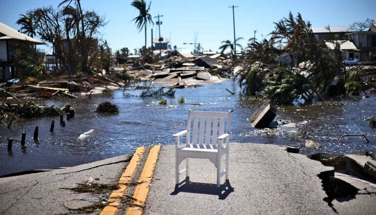 A chair sits at the edge of a destroyed and flooded road, with trees, houses, and debris in the background.