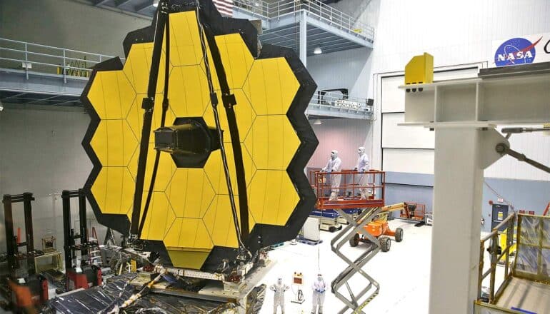 Scientists in white clean suits work on the gigantic James Webb Telescope, one piece of which looks like a honeycomb pattern.