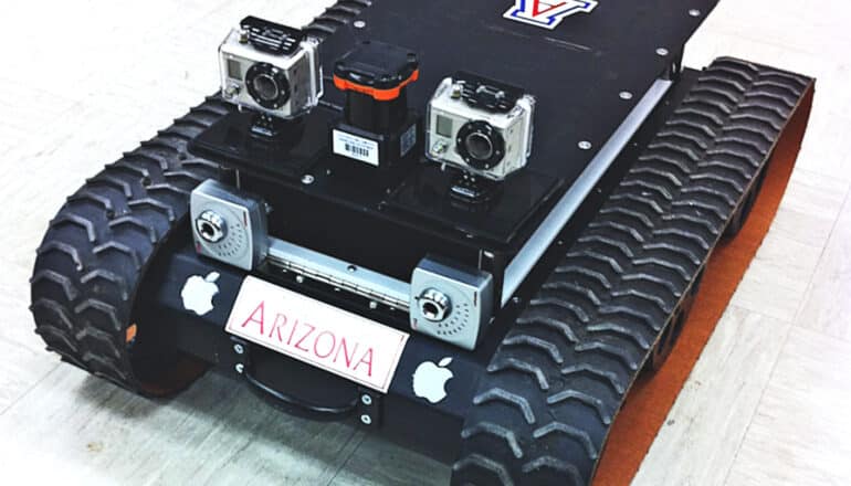 A rover with tank-like treads and equipped with several cameras sits on an office floor.