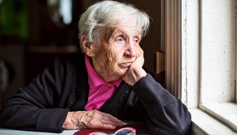 An older woman sits at a kitchen table and rests her head on her hand.