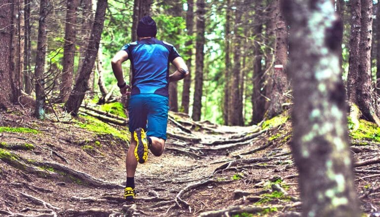 A man in blue workout gear runs along a forest path covered in roots.