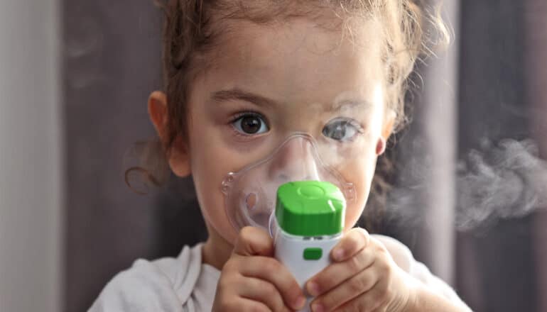 A young girl uses a nebulizer while looking at the camera.