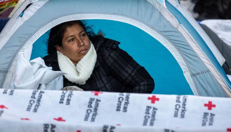person in scarf and coat sits in tent with Red Cross blanket across opening