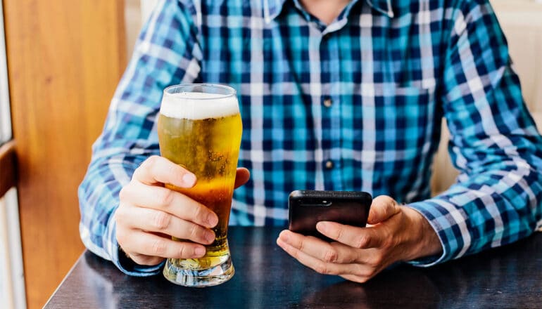 A man holds a beer while sitting at a table looking at his phone.