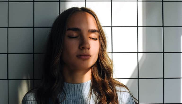 person with closed eyes stands against white tile wall