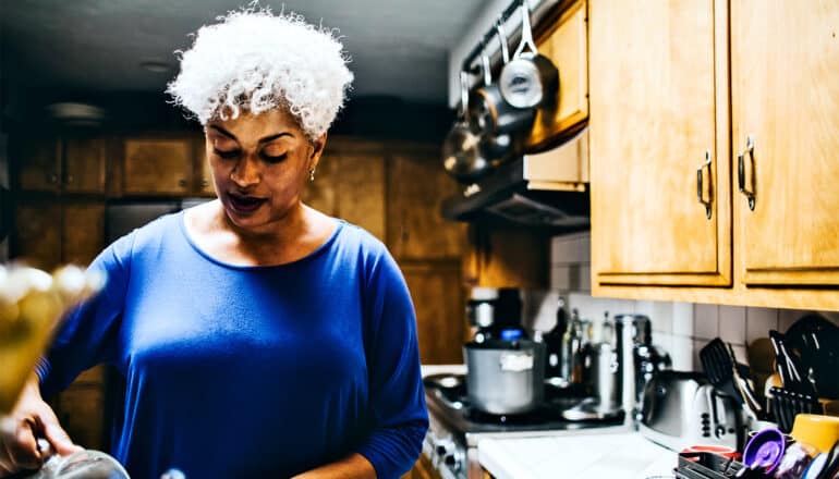 A woman with white hair pours a pitcher of water while standing in a kitchen.