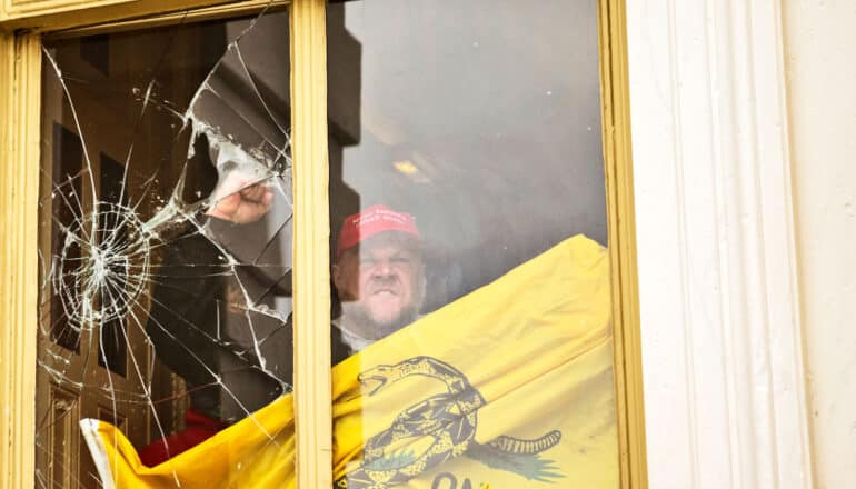 scowling person in MAGA cap holding Gadsden flag breaks window with fist