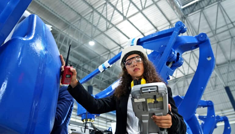 Hispanic woman in hard hat holds clipboard and walkie talkie in front of large blue robot arms