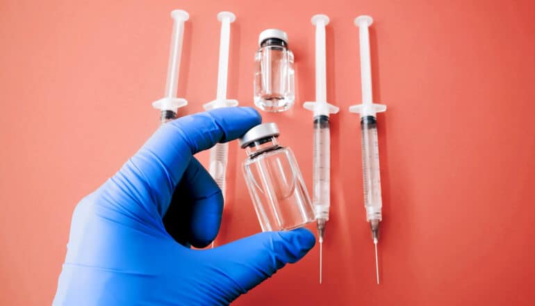 gloved hand holds vial above syringes on tomato red surface