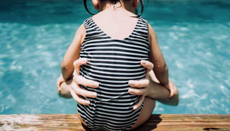 adult hands hold waist of toddler in striped swimsuit who sits on edge of pool