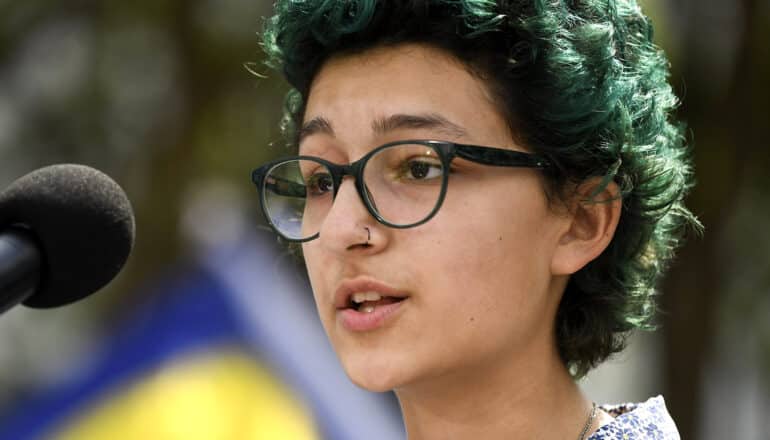 young person with glasses and short green hair speaks into microphone