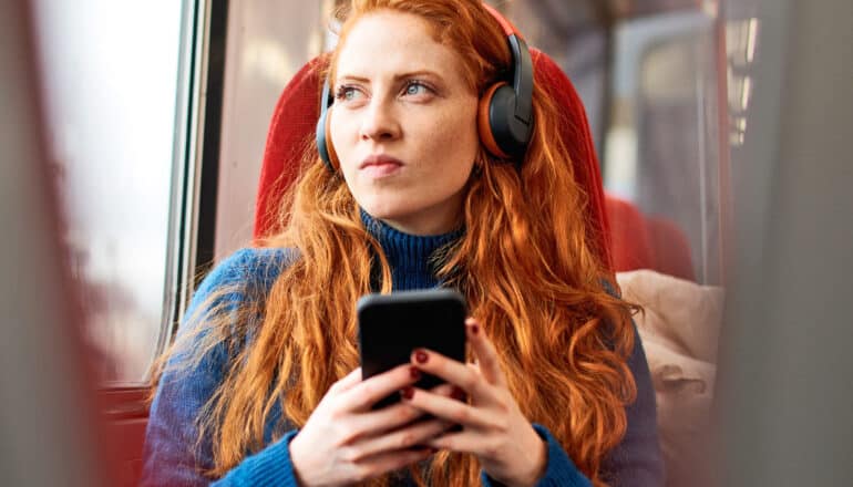 A young woman with bright red hair listens to a podcast while holding her phone and sitting in a train seat.