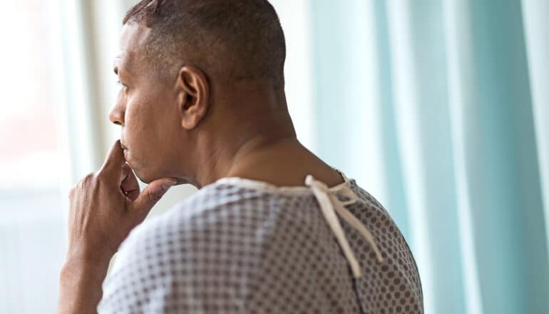 A man in a hospital gown sits looking out a window with his hand on his chin.