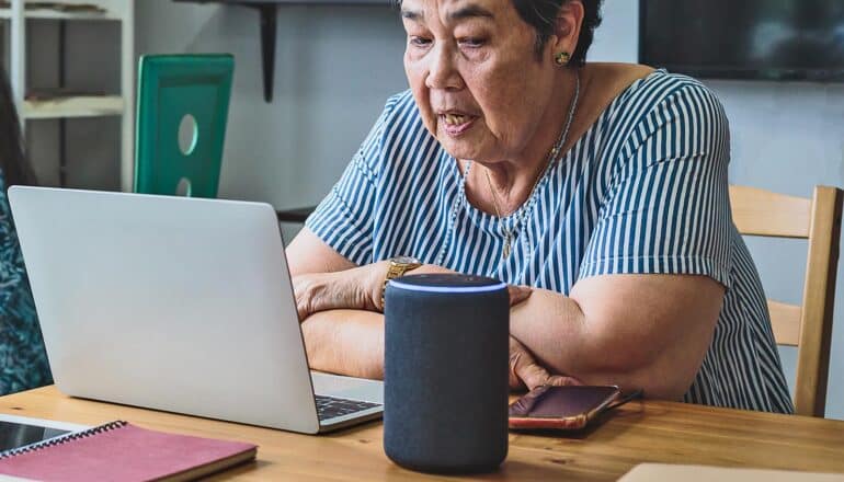 An older woman sitting at a table with a laptop looks down at her voice assistant speaker.