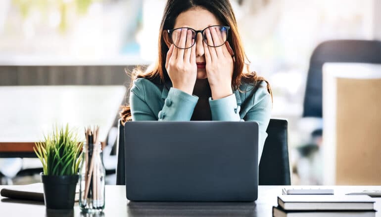 A woman covers her eyes with her hands under her glasses while sitting at her laptop.