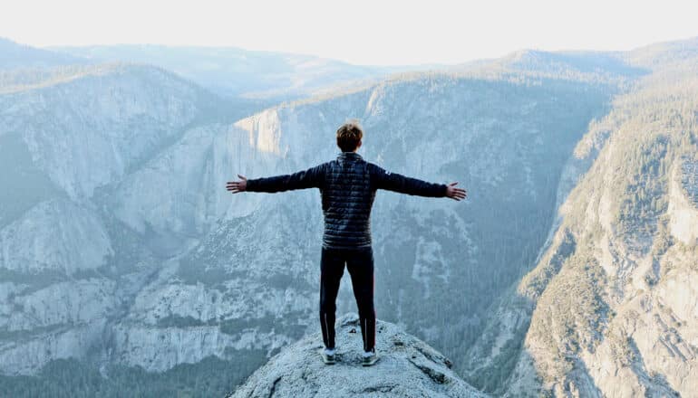 person stands on mountain cliff with arms outstretched