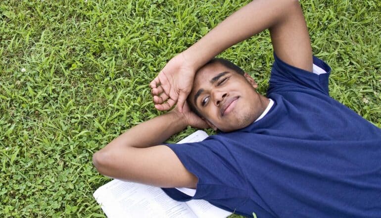 teen lies on book on grass, squints grumpily