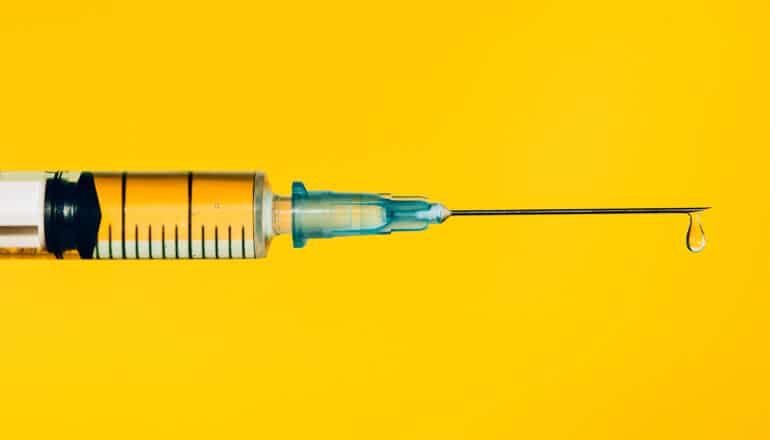 A syringe with liquid coming out of the needle against a yellow background.