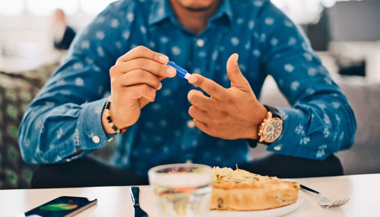 A man in a blue shirt tests his blood sugar while sitting at a desk with food on it.