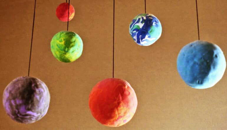 Clay models of planets hang on string against a brown wall.