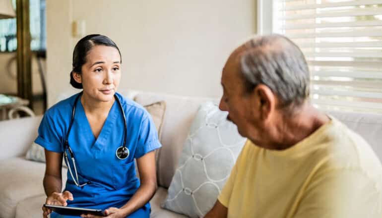 home health nurse uses tablet while sitting on sofa with older adult