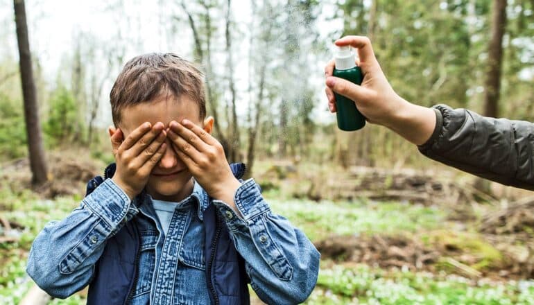 A young boy covers his eyes as a parent sprays him with mosquito repellent.