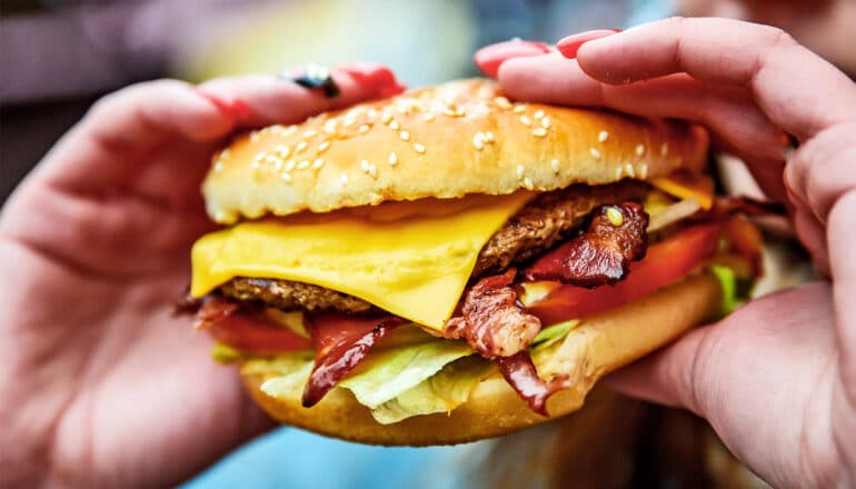 A person with long colorful nails holds a cheeseburger.