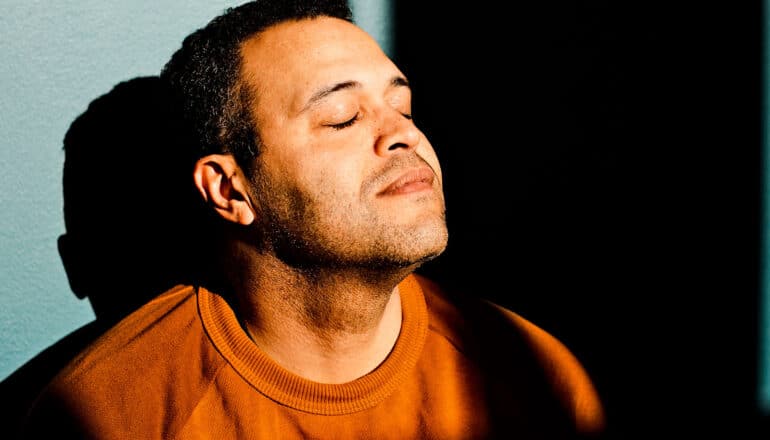 A man sits meditating with his eyes closed against a blue wall.