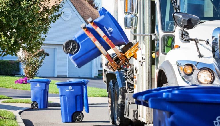 A recycling truck lifts a blue bin to empty it into the truck.