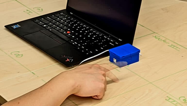 A laptop on a desk has the sensor system hooked up to it while a person uses the desk its sitting on as a touch screen.