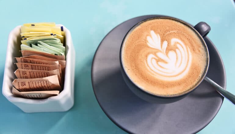 A cappuccino on a blue table next to some sugar and artificial sweetener packets.