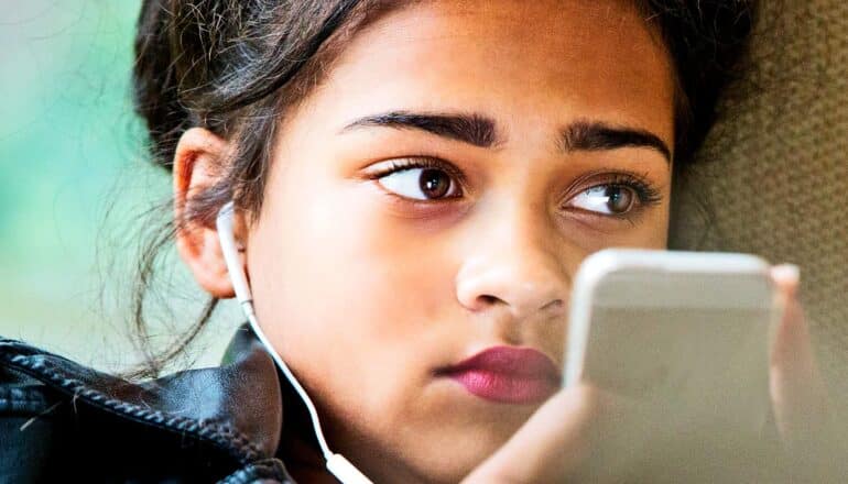 A teen girl looks off camera while holding her phone and wearing headphones.