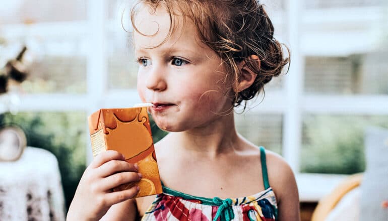 A young girl drinks from a juice box while standing outside.