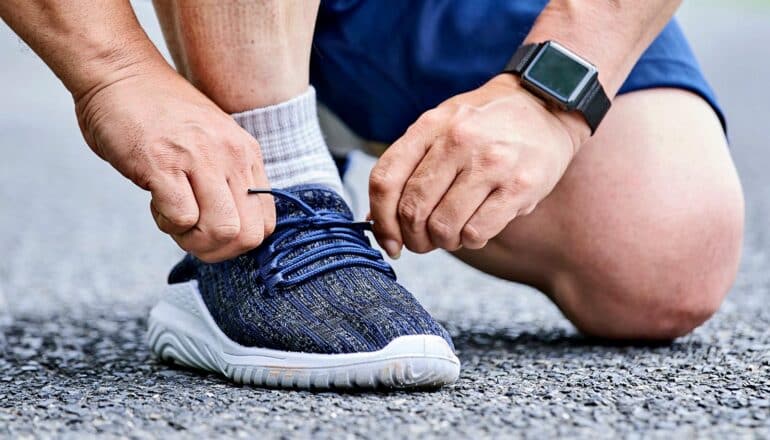 A man wearing an activity tracker ties his running shoes while kneeling on asphalt.