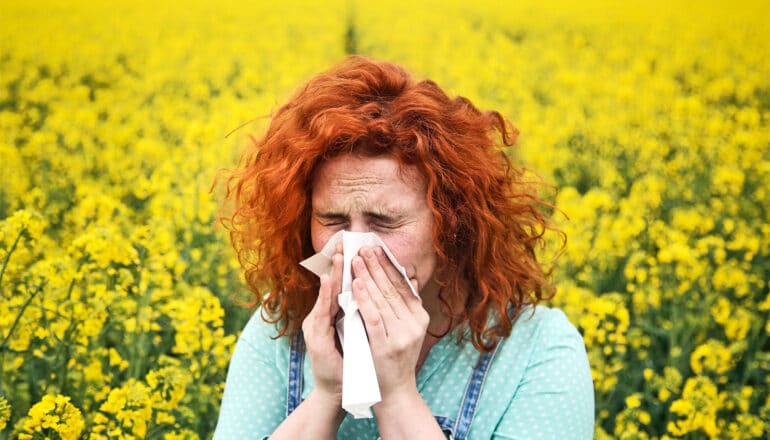 A woman with bright red hair stands in a field of yellow flowers blowing her nose.