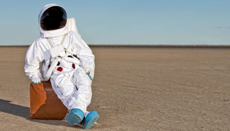 An astronaut sits on a suitcase in the middle of an otherworldly desert.