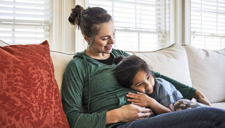 pregnant adult sits on sofa with young child lying across lap