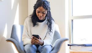 A young woman sits in a large library chair and looks down at her phone.