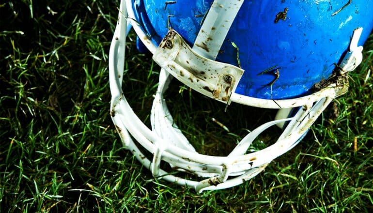 A blue football helmet splattered with grass and mud sits on a green field.