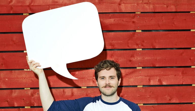 A man holds a white speech bubble cut out of cardboard up over his head as he stands in front of a red wood wall.