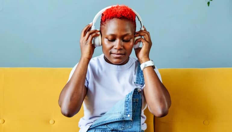 A woman puts on headphones to listen to music while sitting on a couch.