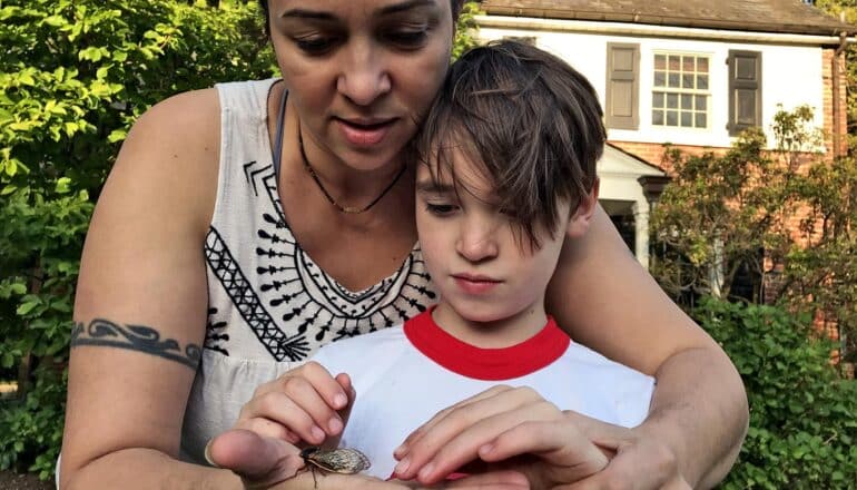 adult with arms around child holds cicada on hand